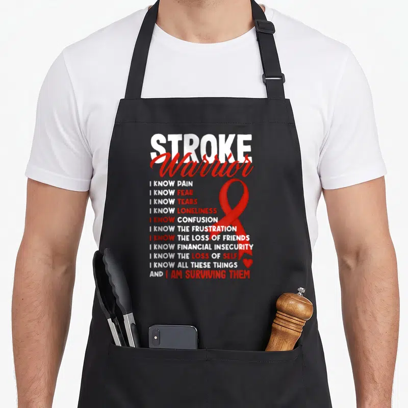 Warrior survivor stroke awareness apron - STROKE Warrior text with red ribbon and list of survival experiences on black — shown on a black cotton canvas apron against a white background