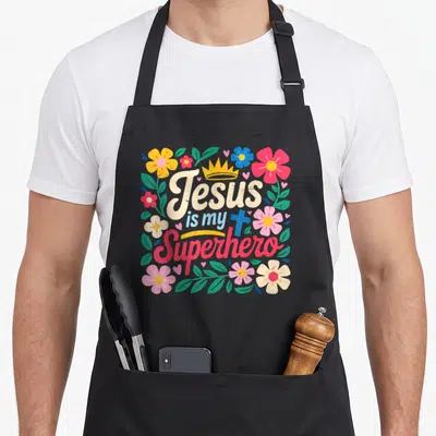 Sister christian apron - Stacked text Jesus is my Superhero surrounded by colorful flowers and hearts — shown on a black cotton canvas apron against a white background