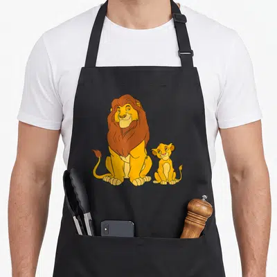 Mufasa lion apron - Cartoon adult lion and cub sitting side by side in yellow and brown — shown on a black cotton canvas apron against a white background