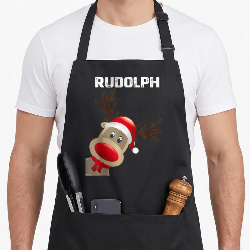 Rudolph ugly christmas apron - Cartoon reindeer wearing red hat and scarf with brown antlers and white RUDOLPH text — shown on a black cotton canvas apron against a white background