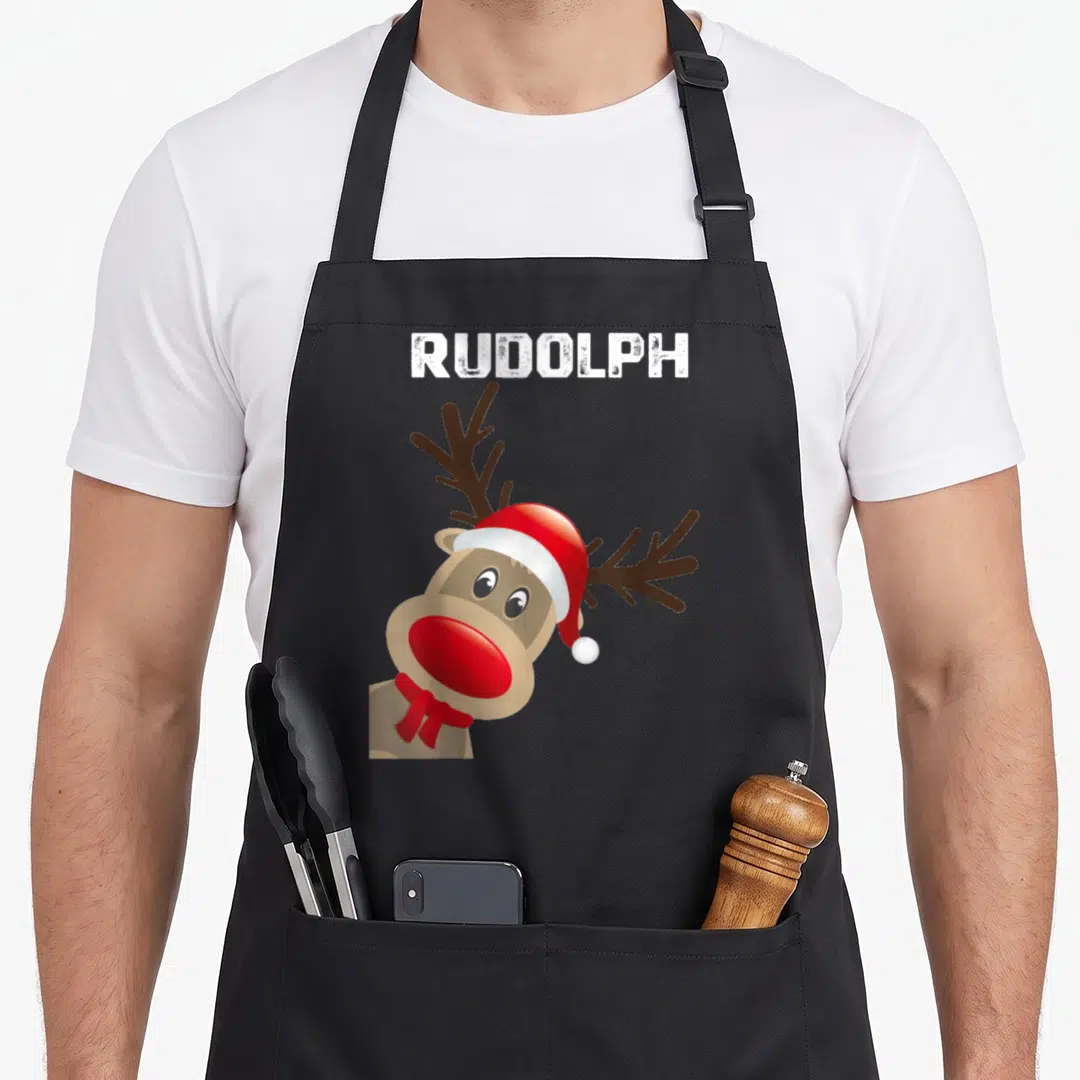 Rudolph ugly christmas apron - Cartoon reindeer wearing red hat and scarf with brown antlers and white RUDOLPH text — shown on a black cotton canvas apron against a white background