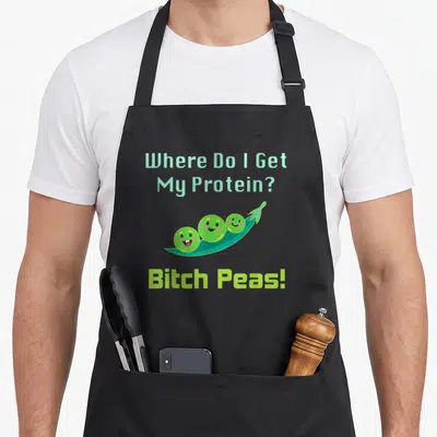 Protein vegan apron - Three smiling peas in teal pod with lime green Bitch Peas text — shown on a black cotton canvas apron against a white background