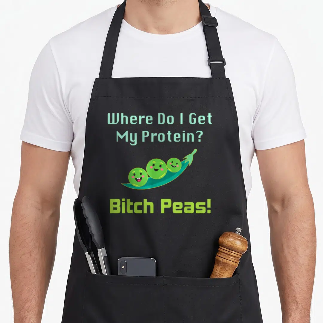 Protein vegan apron - Three smiling peas in teal pod with lime green Bitch Peas text — shown on a black cotton canvas apron against a white background