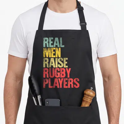 Raise rugby life apron - Stacked text REAL MEN RAISE RUGBY PLAYERS in green yellow and red distressed typography — shown on a black cotton canvas apron against a white background