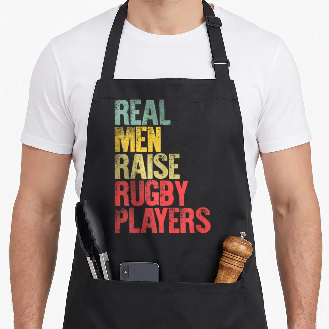 Raise rugby life apron - Stacked text REAL MEN RAISE RUGBY PLAYERS in green yellow and red distressed typography — shown on a black cotton canvas apron against a white background