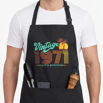 50th aged birthday apron - Retro 1971 text with palm trees and sun in teal orange yellow — shown on a black cotton canvas apron against a white background