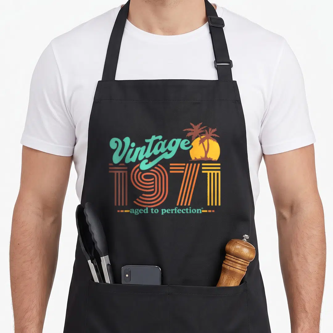 50th aged birthday apron - Retro 1971 text with palm trees and sun in teal orange yellow — shown on a black cotton canvas apron against a white background