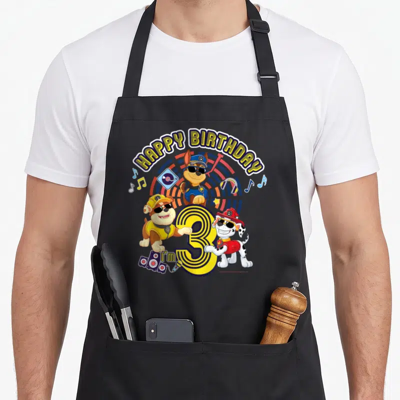 Funny age birthday apron - Three cartoon puppies with sunglasses surround yellow number 3 and I'm 3 text with music — shown on a black cotton canvas apron against a white background