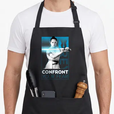 Rise fear apron - Confront Your Fear text below grayscale woman holding blue lightsaber on blue background — shown on a black cotton canvas apron against a white background