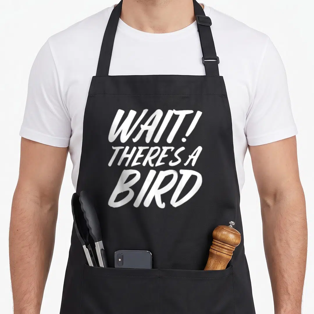Watcher birding bird apron - White stacked text WAIT THERES A BIRD on grey background — shown on a black cotton canvas apron against a white background