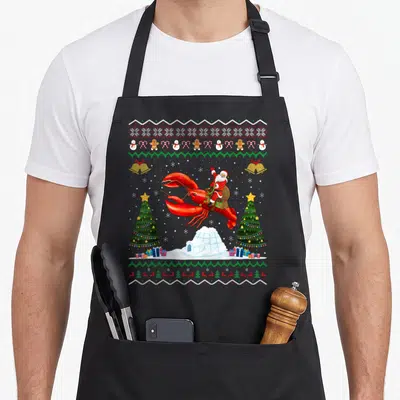Lobster xmas apron - Santa riding red lobster with Christmas trees, igloo, and pixelated knit border in red green white — shown on a black cotton canvas apron against a white background