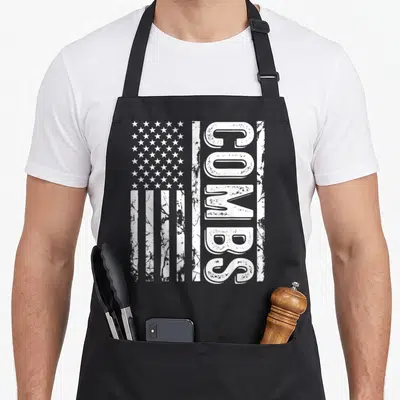 Comb family reunion aprons - Vertical white USA flag with distressed COMBS text running alongside stars and stripes — shown on a black cotton canvas apron against a white background