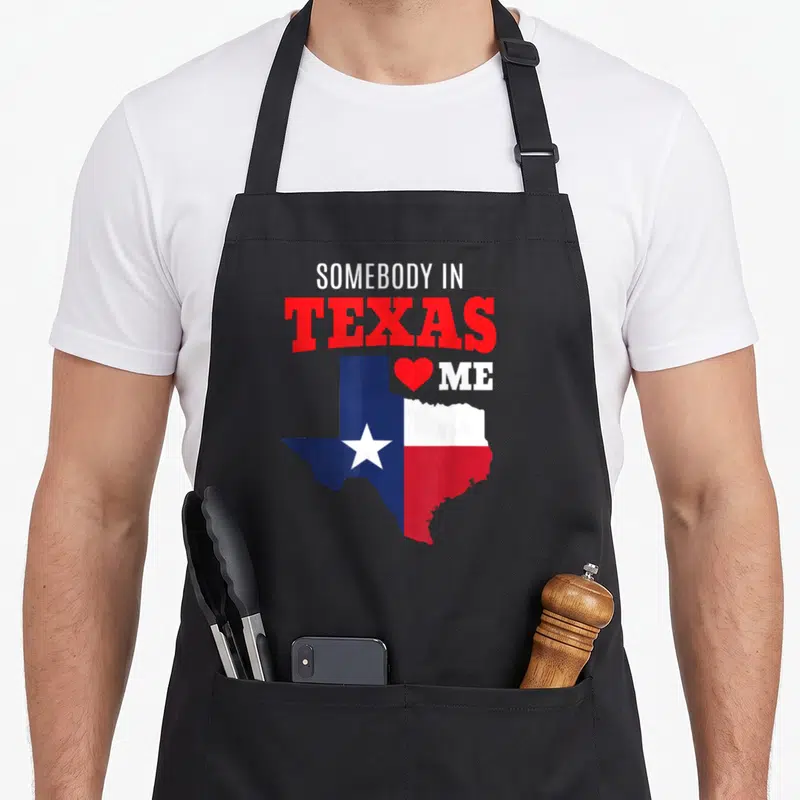 Somebody texas apron - Stacked text SOMEBODY IN TEXAS ME with red heart above Texas map in flag colors — shown on a black cotton canvas apron against a white background