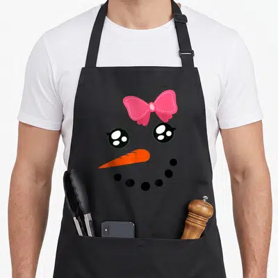 Snow bow apron - Cartoon snowman face with pink bow, two black eyes, orange carrot nose, and curved row of black dots — shown on a black cotton canvas apron against a white background