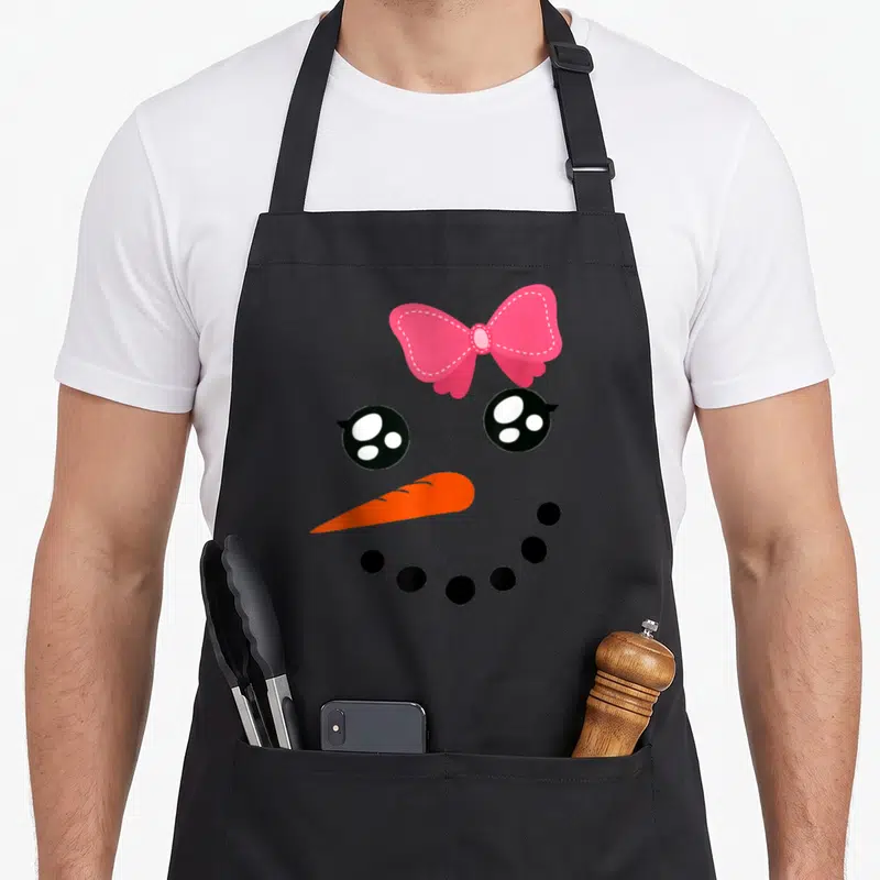 Snow bow apron - Cartoon snowman face with pink bow, two black eyes, orange carrot nose, and curved row of black dots — shown on a black cotton canvas apron against a white background