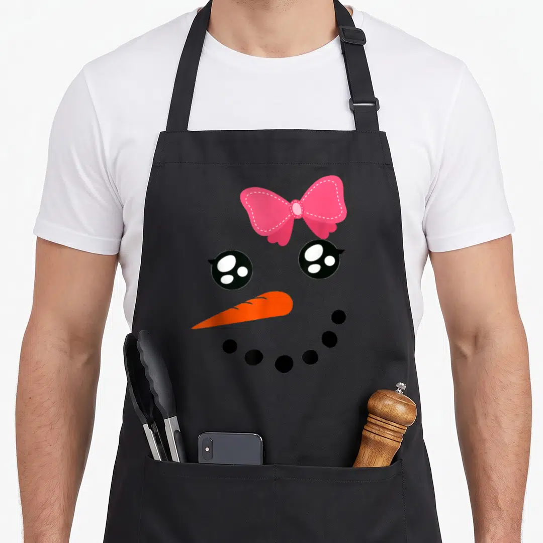 Snow bow apron - Cartoon snowman face with pink bow, two black eyes, orange carrot nose, and curved row of black dots — shown on a black cotton canvas apron against a white background