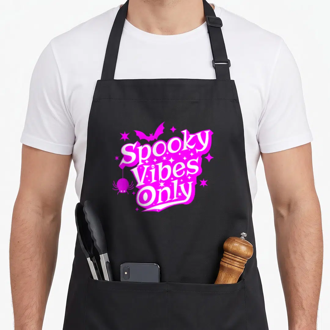 Funny spooky halloween apron - Spooky Vibes Only text with pink bat, spider, skull, and stars on pink shape — shown on a black cotton canvas apron against a white background