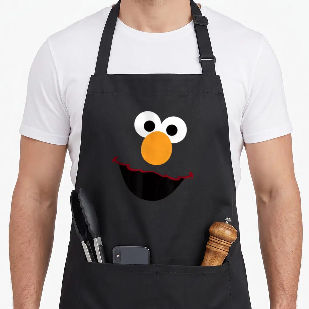 Elmo face halloween apron - Red cartoon face with white eyes orange nose and black smiling mouth — shown on a black cotton canvas apron against a white background