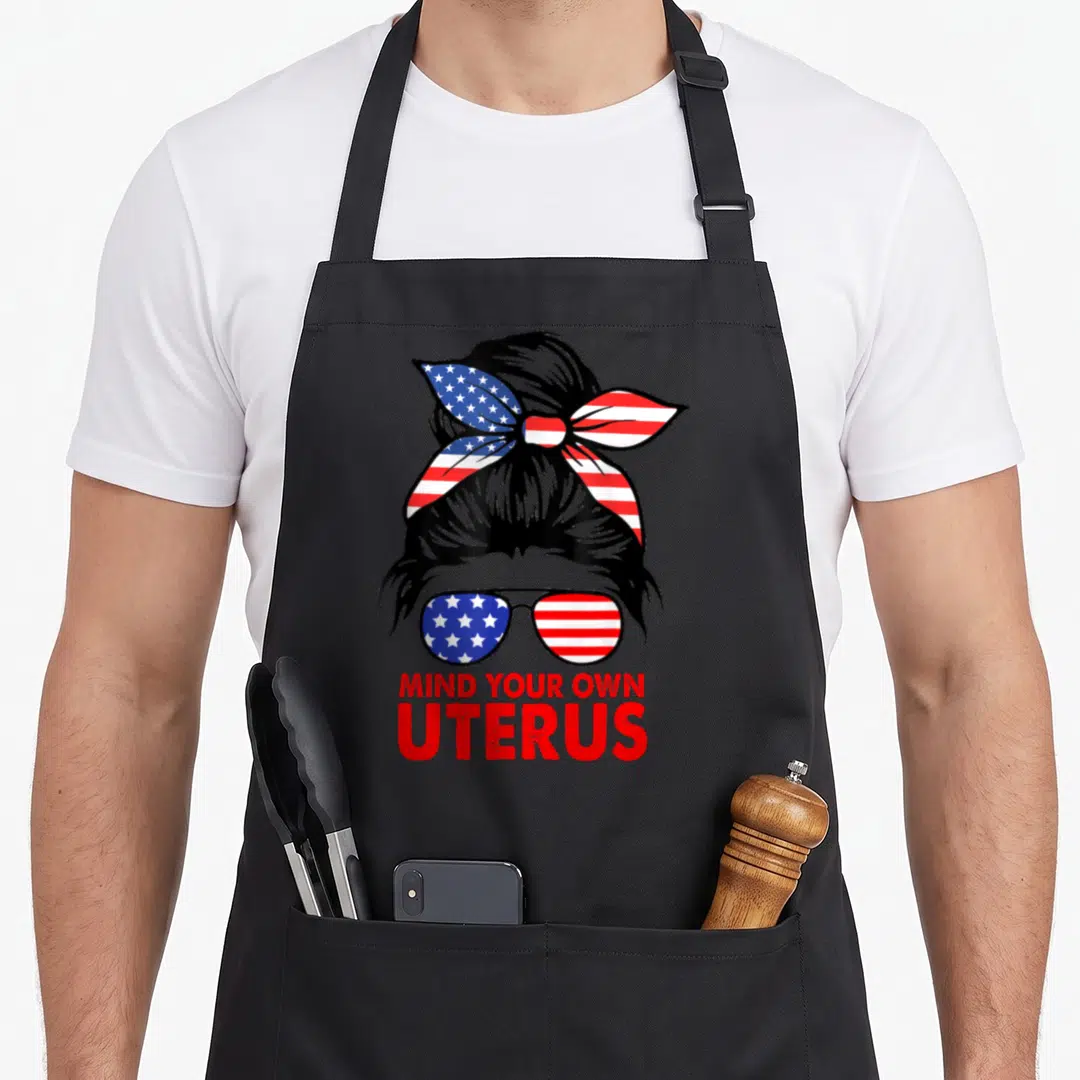 Flag messy aprons - Woman silhouette with US flag bow and sunglasses above red text Mind Your Own Uterus — shown on a black cotton canvas apron against a white background