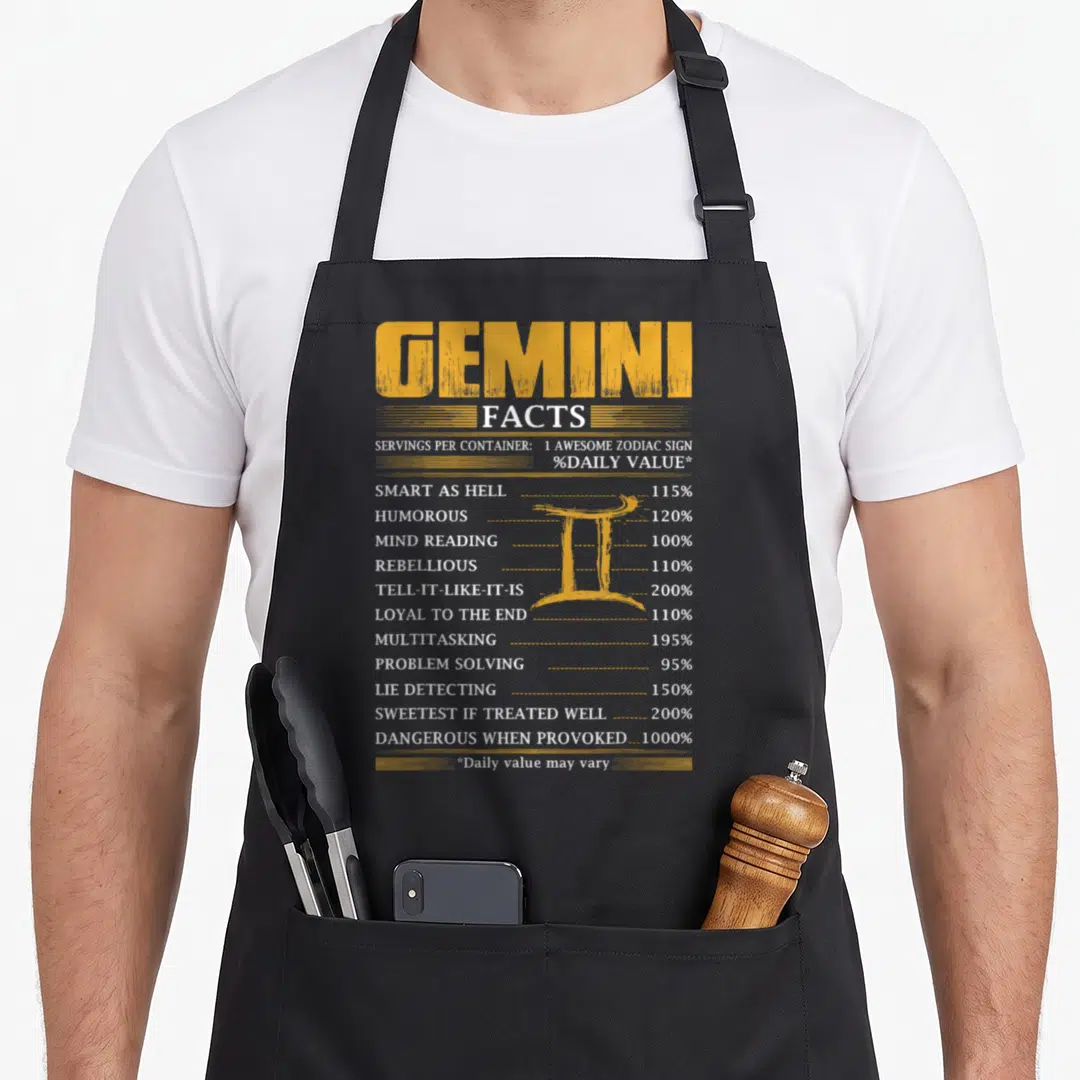 Gemini serving apron - Distressed yellow nutrition label listing Gemini traits like rebellious and loyal with a central — shown on a black cotton canvas apron against a white background