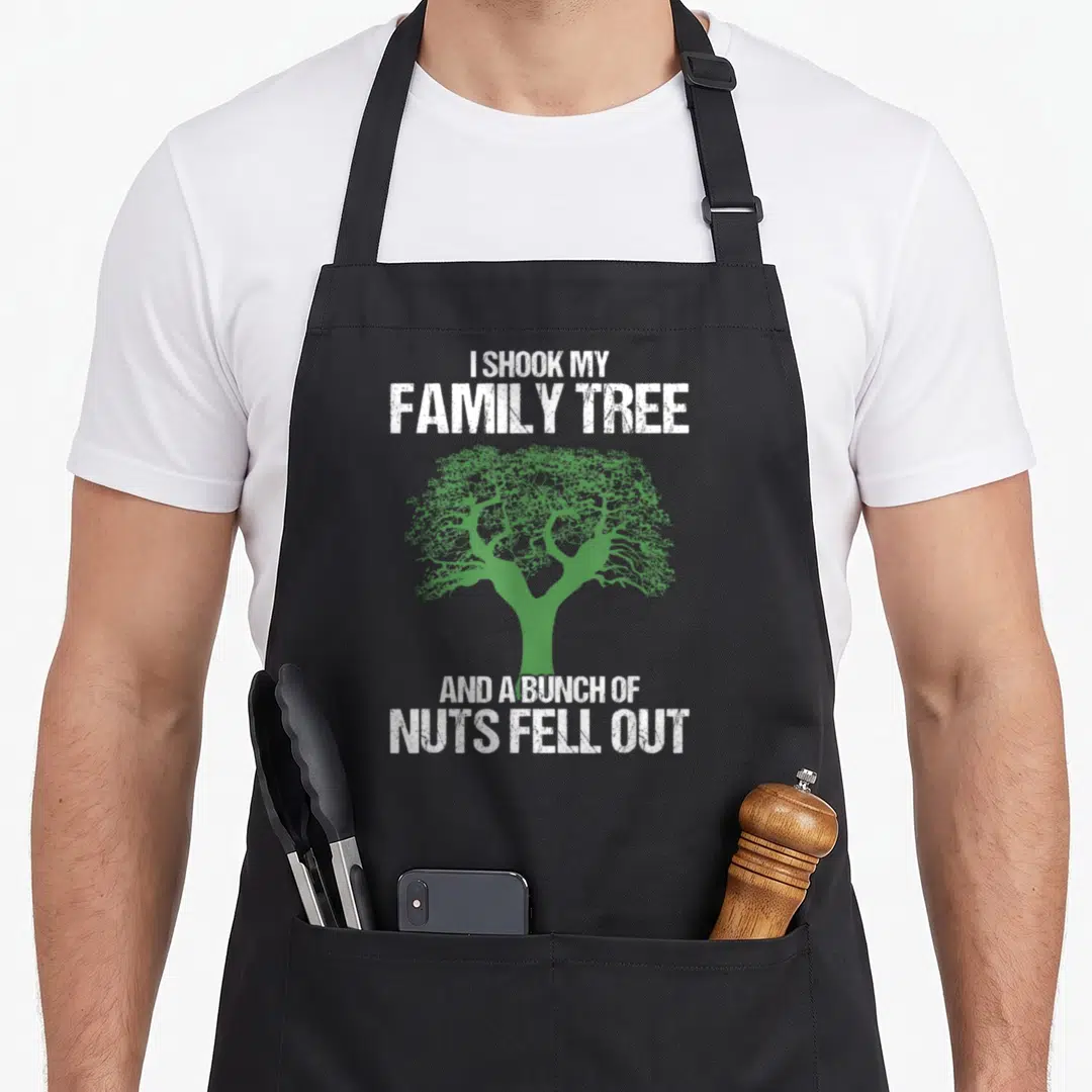 Genealogie tree apron - Distressed white text I SHOOK MY FAMILY TREE AND A BUNCH OF NUTS FELL OUT around a green tree — shown on a black cotton canvas apron against a white background