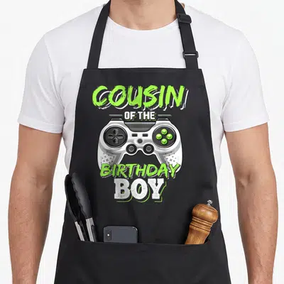 Cousin video game apron - Lime green and white game controller with COUSIN OF THE BIRTHDAY BOY text — shown on a black cotton canvas apron against a white background