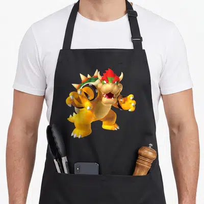 Funny poster holiday apron - 3D cartoon turtle monster with red hair, green shell, and yellow scales centered on fabric — shown on a black cotton canvas apron against a white background