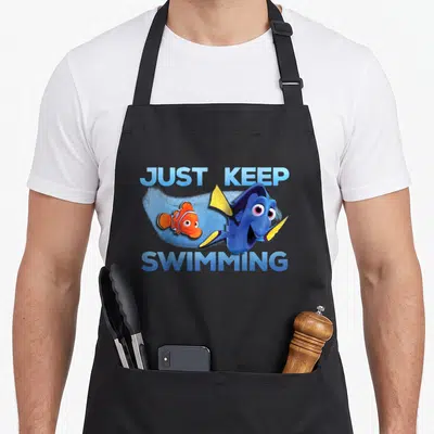 Finding swimming apron - Dory and Nemo swimming under JUST KEEP SWIMMING text in blue orange yellow cartoon style — shown on a black cotton canvas apron against a white background