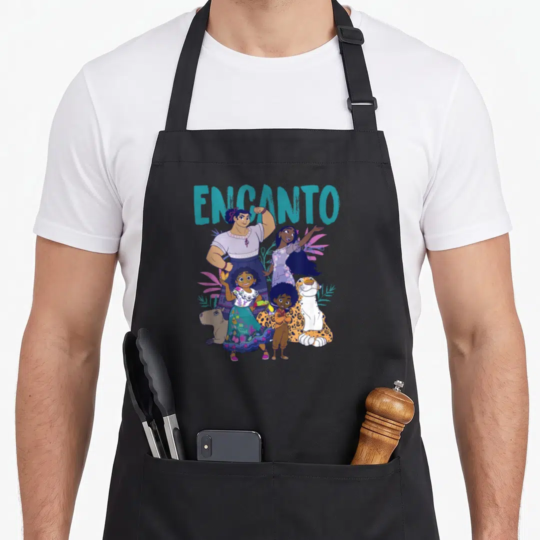Shot encinitas apron - ENCANTO text above cartoon characters, jaguar, capybara, and pink teal leaves — shown on a black cotton canvas apron against a white background