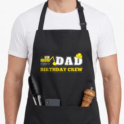 Crew daddy apron - Cartoon excavator and hard hat next to DAD BIRTHDAY CREW text — shown on a black cotton canvas apron against a white background