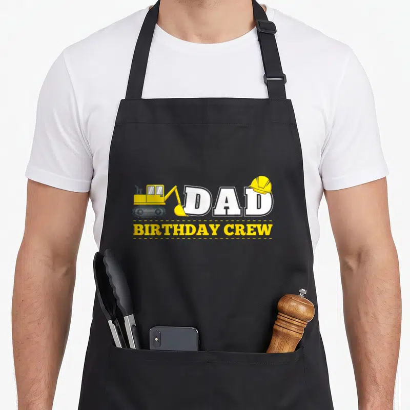 Crew daddy apron - Cartoon excavator and hard hat next to DAD BIRTHDAY CREW text — shown on a black cotton canvas apron against a white background