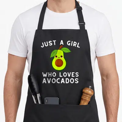 Guacamole avocado apron - Cartoon avocado with green leaves and brown pit under white text reading JUST A GIRL WHO — shown on a black cotton canvas apron against a white background