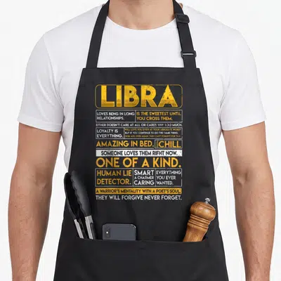 Libra horoscope apron - Yellow and white text blocks listing Libra traits like loyalty and human lie detector on black — shown on a black cotton canvas apron against a white background