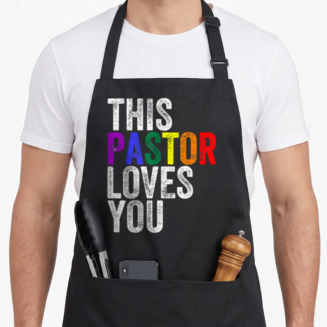 Pastor gay pride apron - Stacked text THIS PASTOR LOVES YOU with rainbow colored PASTOR on black background — shown on a black cotton canvas apron against a white background