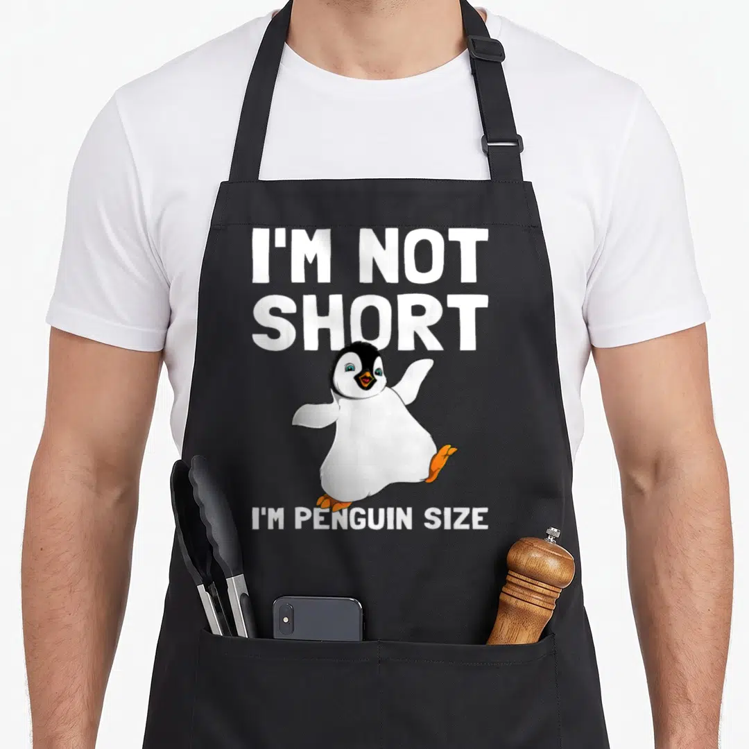 Bird penguin aprons - Cartoon penguin with text I'm Not Short I'm Penguin Size in white and orange — shown on a black cotton canvas apron against a white background
