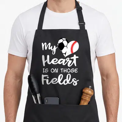 Those baseball apron - Heart shape from soccer ball and baseball with white text My Heart Is On Those Fields — shown on a black cotton canvas apron against a white background