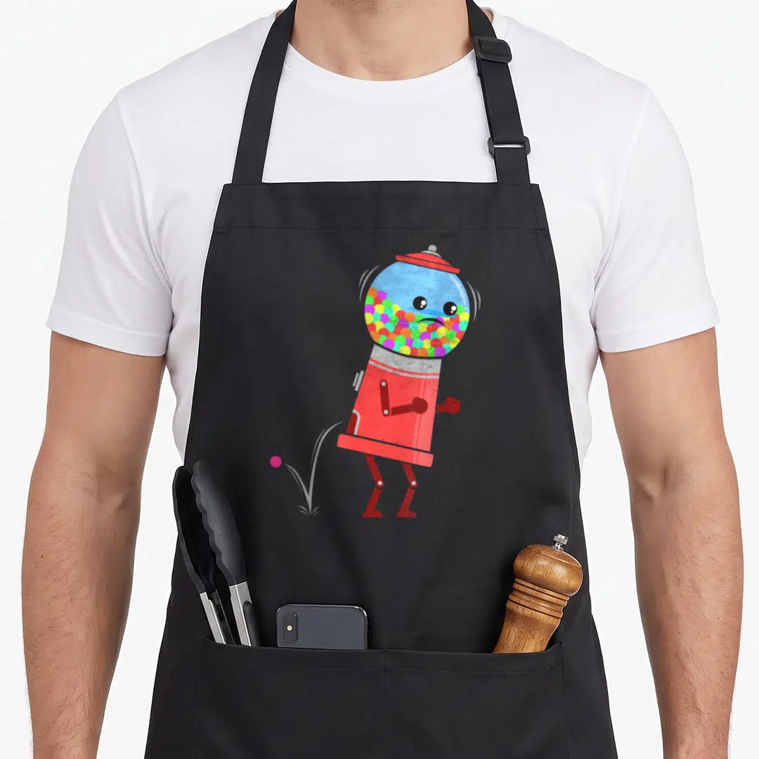 Chewing old fashioned aprons - Cartoon red gumball machine facing left with pink ball on ground and multi-colored balls — shown on a black cotton canvas apron against a white background