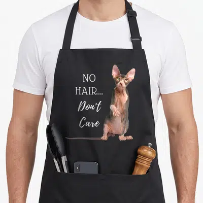 Hair cat apron - No Hair Don't Care text next to a sitting pink and grey hairless cat photo — shown on a black cotton canvas apron against a white background