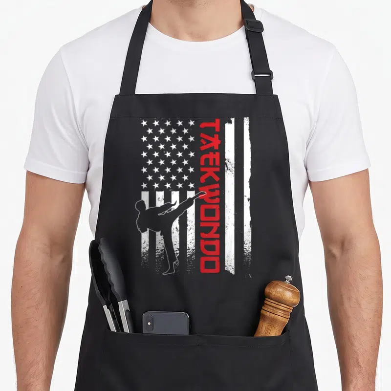 Taekwondo american flag apron - Vertical red TAEKWONDO text beside white kicker silhouette over distressed red — shown on a black cotton canvas apron against a white background