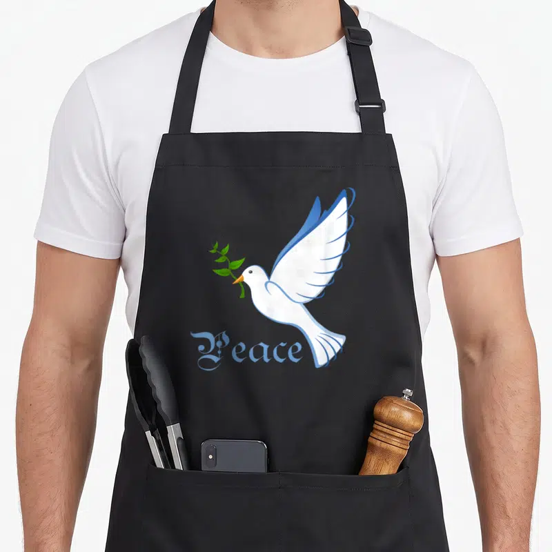 Peace dove apron - White cartoon dove holding green olive branch above blue Peace text — shown on a black cotton canvas apron against a white background
