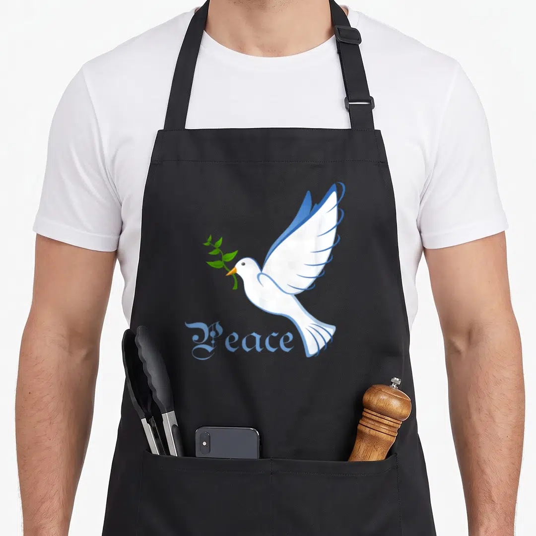 Peace dove apron - White cartoon dove holding green olive branch above blue Peace text — shown on a black cotton canvas apron against a white background