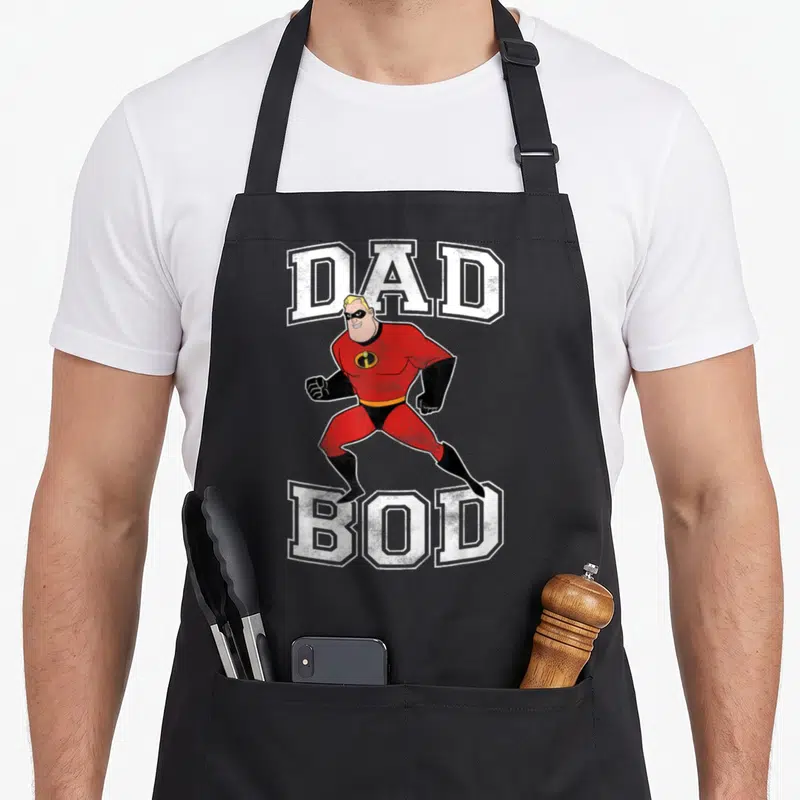 Incredible dad bod apron - Superhero running between DAD and BOD text in red white black — shown on a black cotton canvas apron against a white background