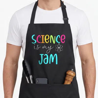 Science jam teacher apron - Multi-colored SCIENCE is my JAM text with white atom symbol on grey background — shown on a black cotton canvas apron against a white background