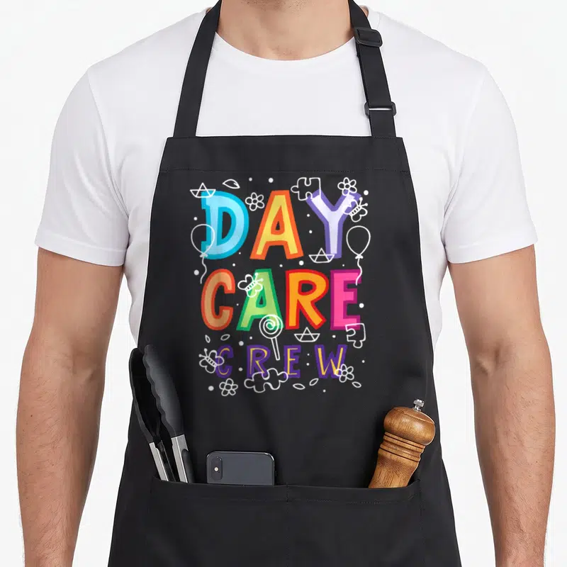 Daycare preschool teacher apron - Stacked DAY CARE CREW text in rainbow colors surrounded by white line art toys — shown on a black cotton canvas apron against a white background