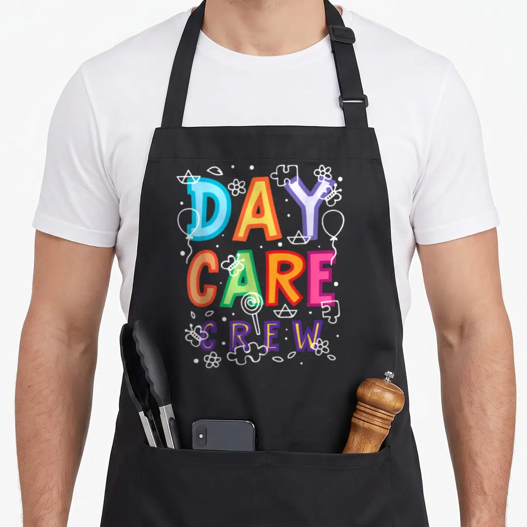 Daycare preschool teacher apron - Stacked DAY CARE CREW text in rainbow colors surrounded by white line art toys — shown on a black cotton canvas apron against a white background