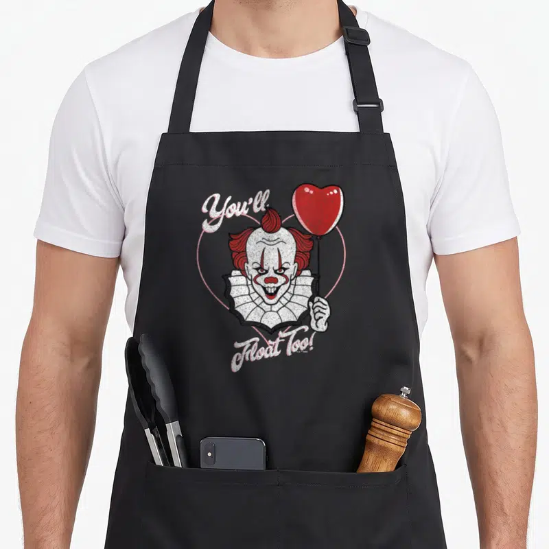 Youll valentine's day apron - Cartoon clown holding red heart balloon with You'll Float Too text inside pink outline — shown on a black cotton canvas apron against a white background