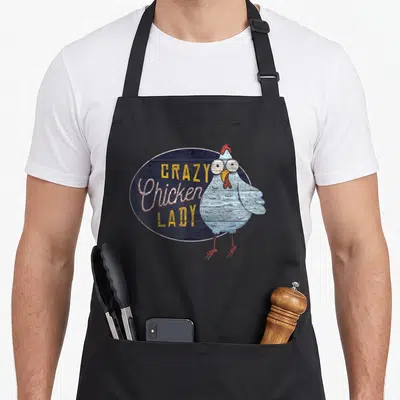 Distressed chicken with apron - Crazy Chicken Lady text next to light blue cartoon hen on dark blue wood oval — shown on a black cotton canvas apron against a white background
