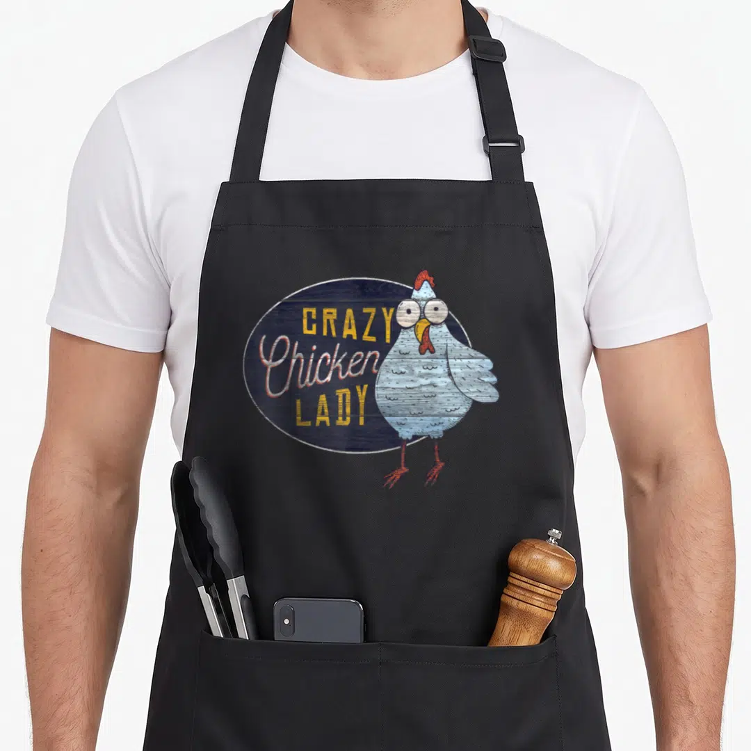 Distressed chicken with apron - Crazy Chicken Lady text next to light blue cartoon hen on dark blue wood oval — shown on a black cotton canvas apron against a white background