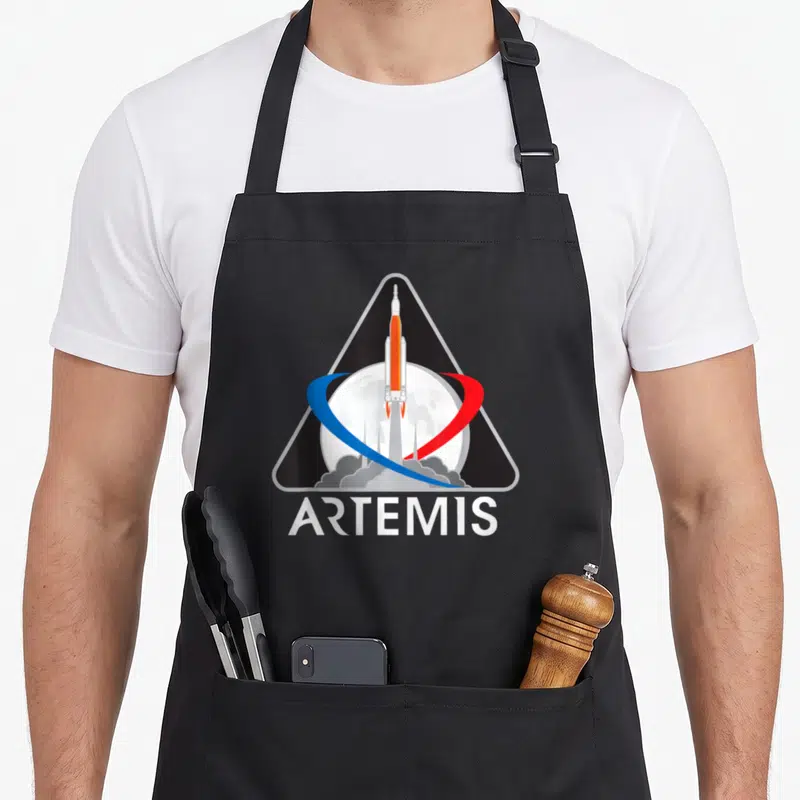 Artemis apollo apron - Rocket and moon inside black triangle with ARTEMIS text and blue red swoosh — shown on a black cotton canvas apron against a white background