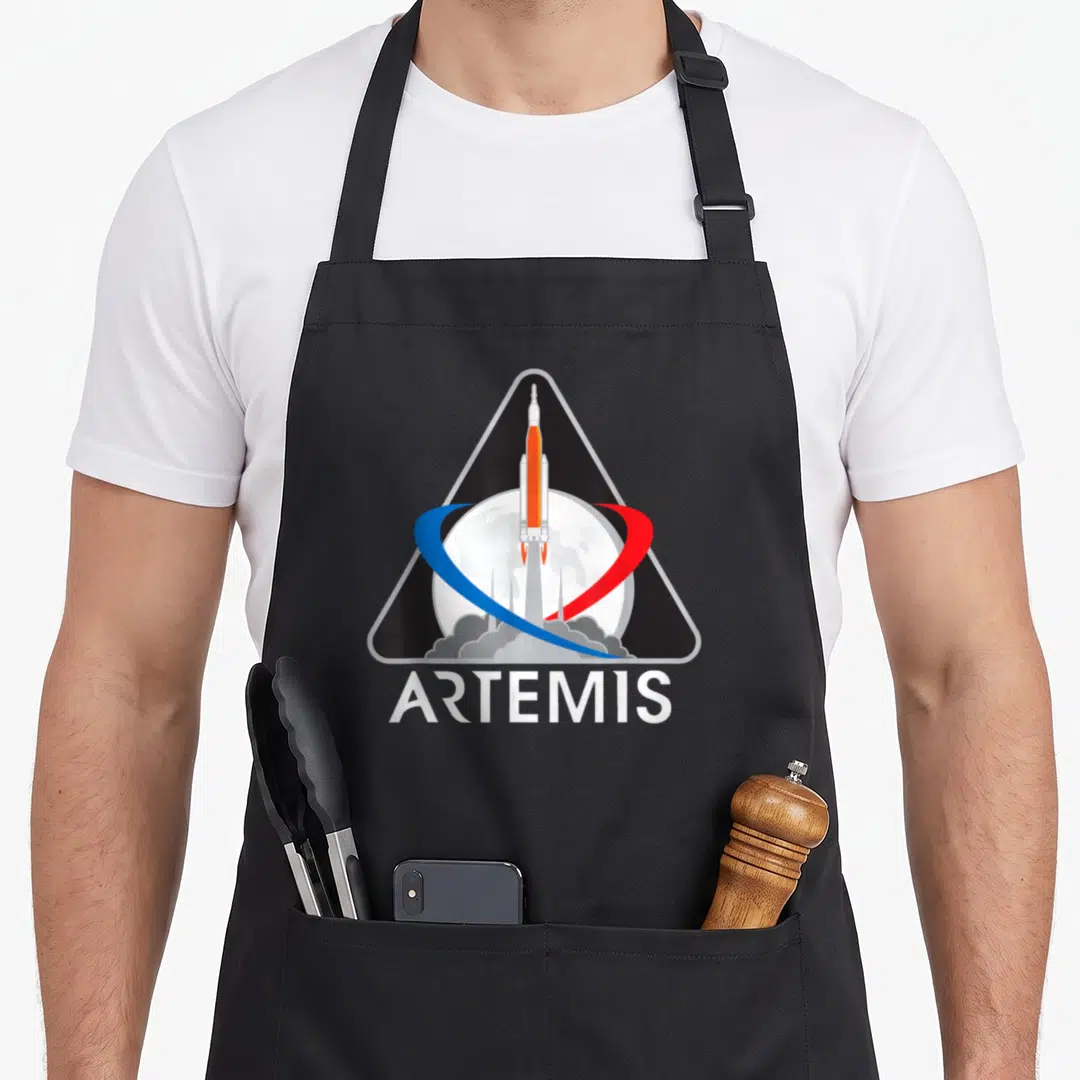 Artemis apollo apron - Rocket and moon inside black triangle with ARTEMIS text and blue red swoosh — shown on a black cotton canvas apron against a white background